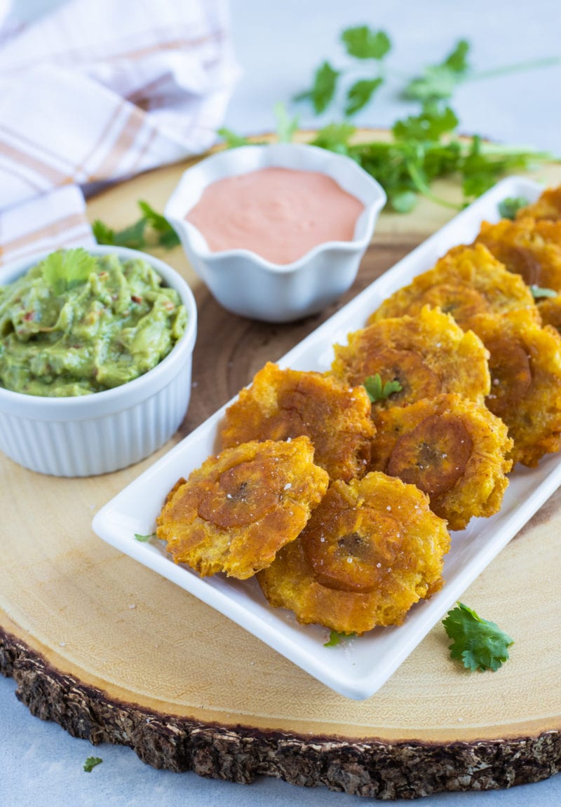 Tostones (Plátanos Verdes Fritos) servidos en un plato blanco con salsa de ketchup de mayonesa y guacamole en el costado.  