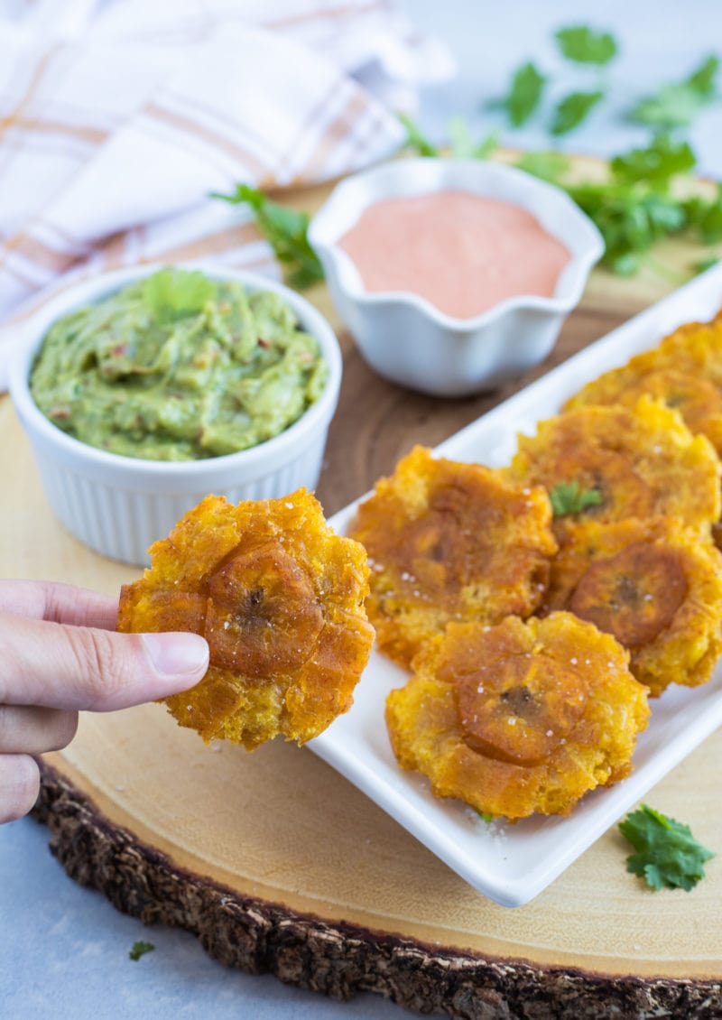 A hand holding a fried tostone next to two bowls of dips.
