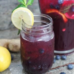 A glass with blueberry lemonade next to a pitcher of the drink.