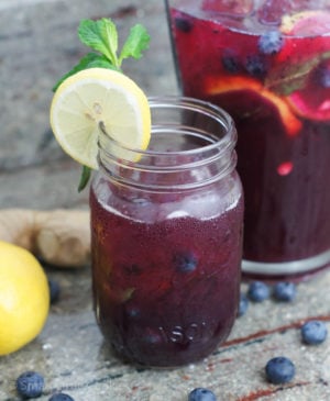 A glass with blueberry lemonade next to a pitcher of the drink.