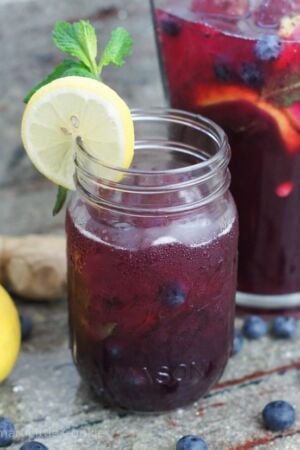 A glass with blueberry lemonade next to a pitcher of the drink.