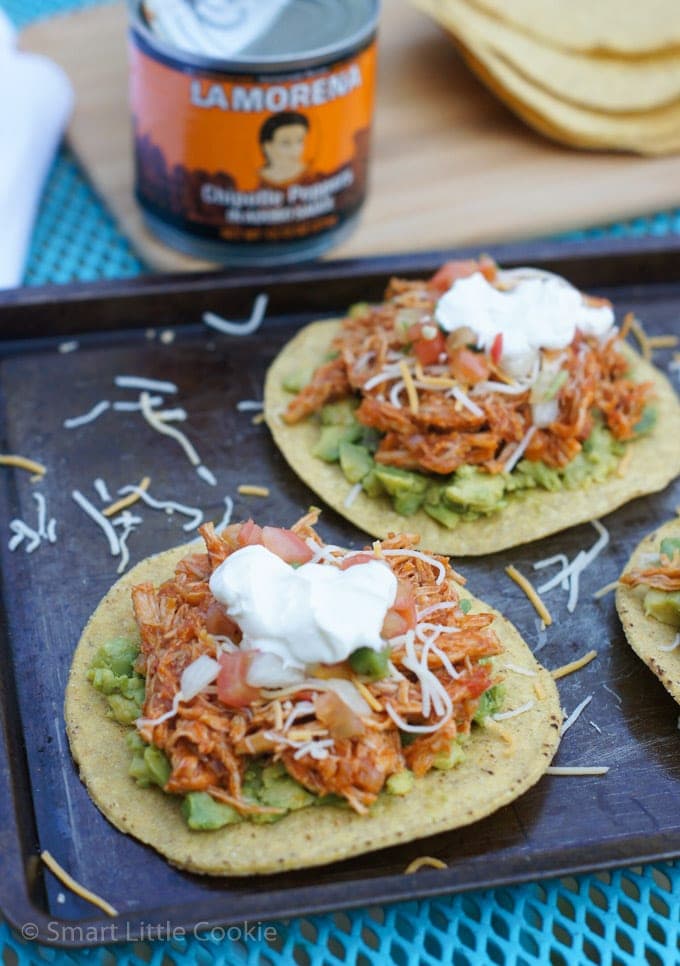 Two chicken tostadas on a baking sheet topped with shredded cheese and sour cream.