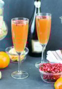 Two tangerine pomegranate mimosas served in flue glasses next to a bowl of pomegranate seeds.
