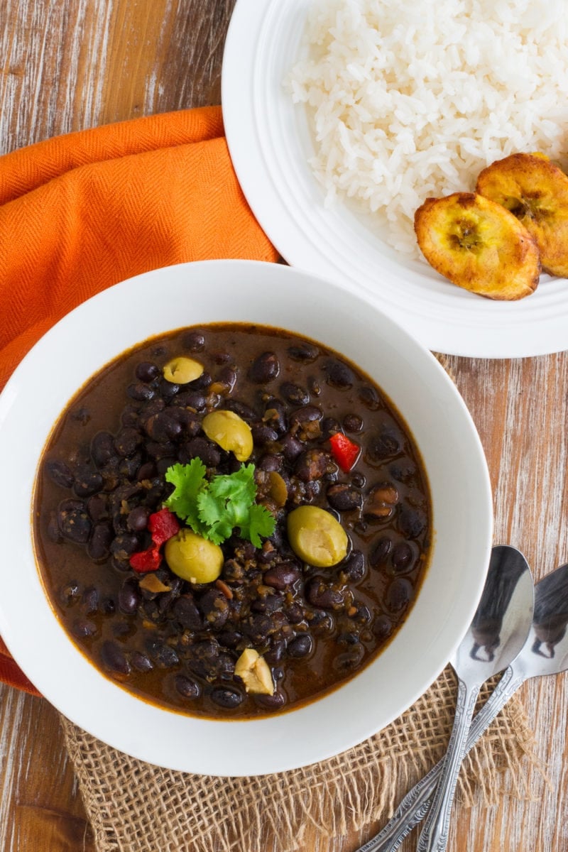 Frijoles Negros guisados servido en un tazón blanco con arroz y plátanos en el lado