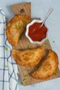 Three chicken parmesan empanadas on a wooden board with a red sauce.