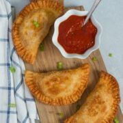 Three chicken parmesan empanadas on a wooden board with a red sauce.