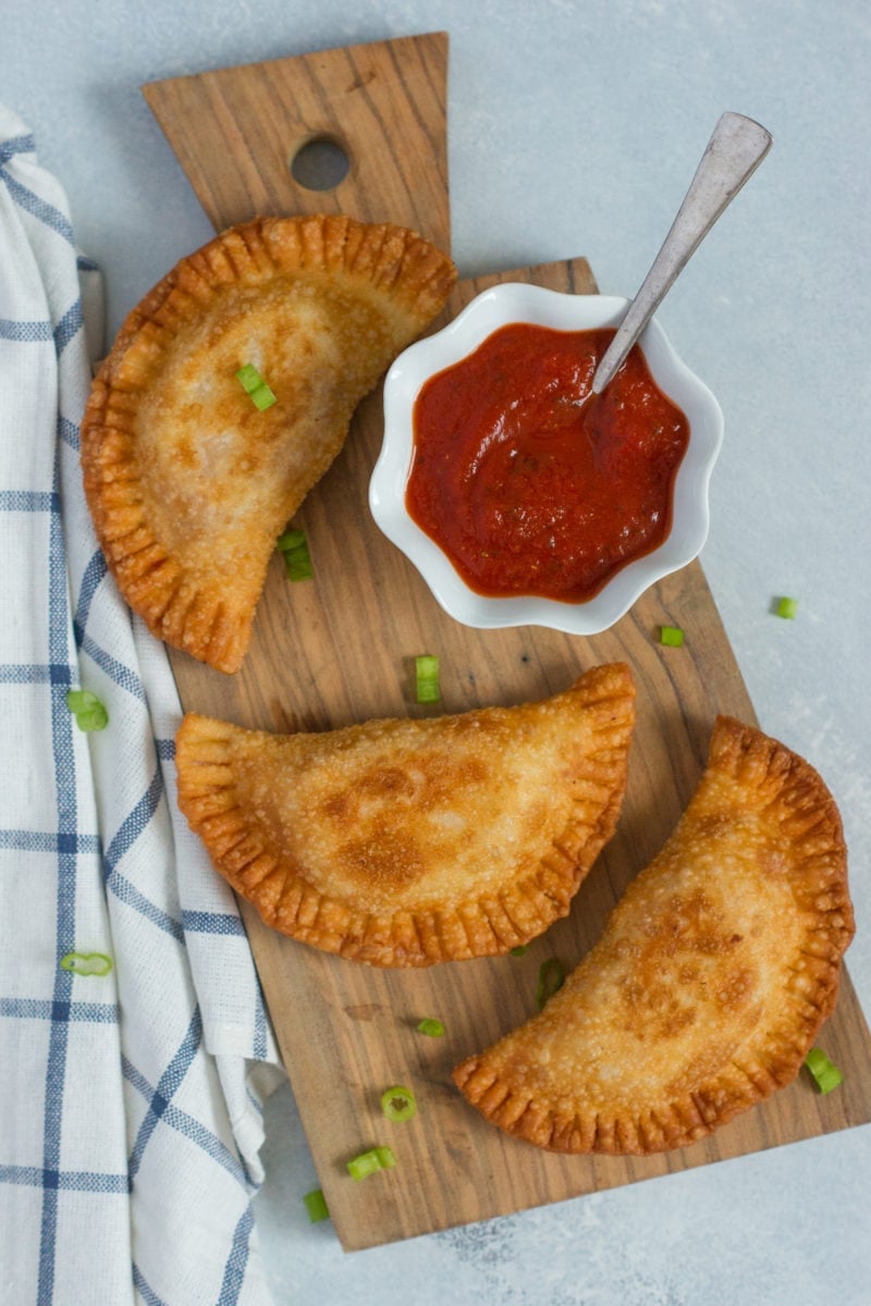 Three chicken parmesan empanadas on a wooden board with a red sauce.