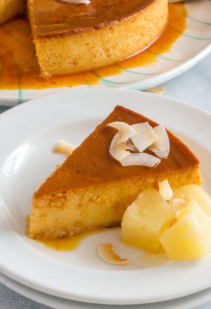 A slice of flan on a white plate next to cubes of pineapple.