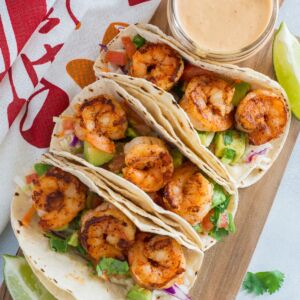 Three shrimp tacos next to a jar of sauce on a wooden chopping board.