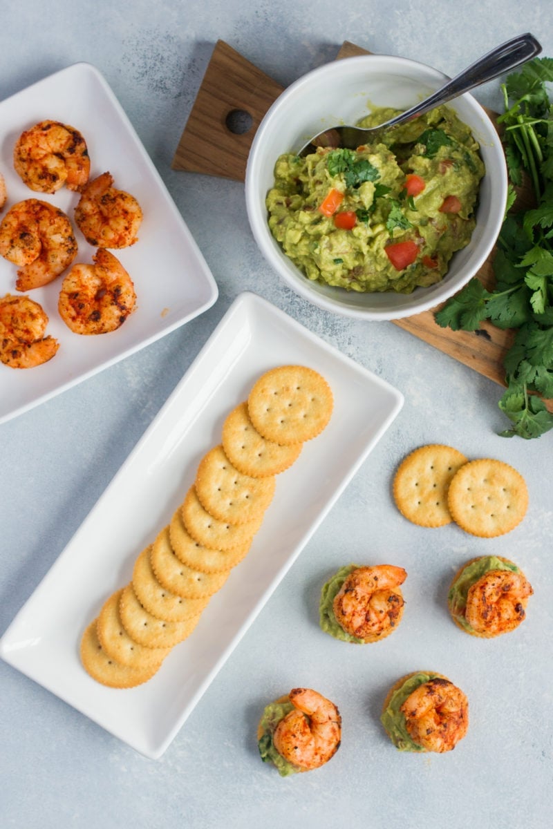 Crackers on a serving plate next to a bowl of guacamole and a plate of shrimp.