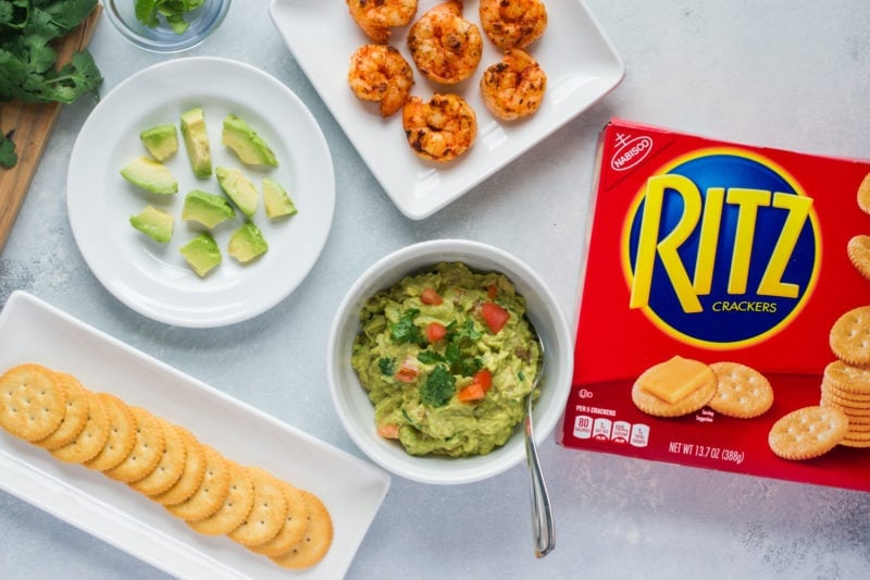 Crackers on a serving plate next to a bowl of guacamole, a plate of shrimp and a box of Ritz crackers.