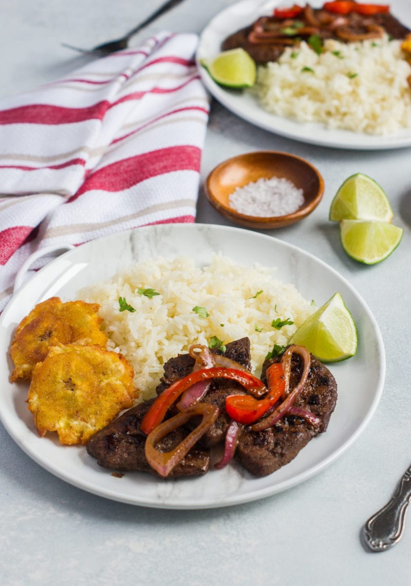 Liver an onions served on a plate with rice and tostones.