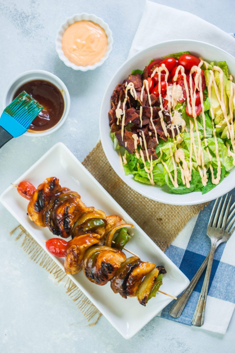 Chicken skewers on a plate next to bbq sauce and a salad bowl.