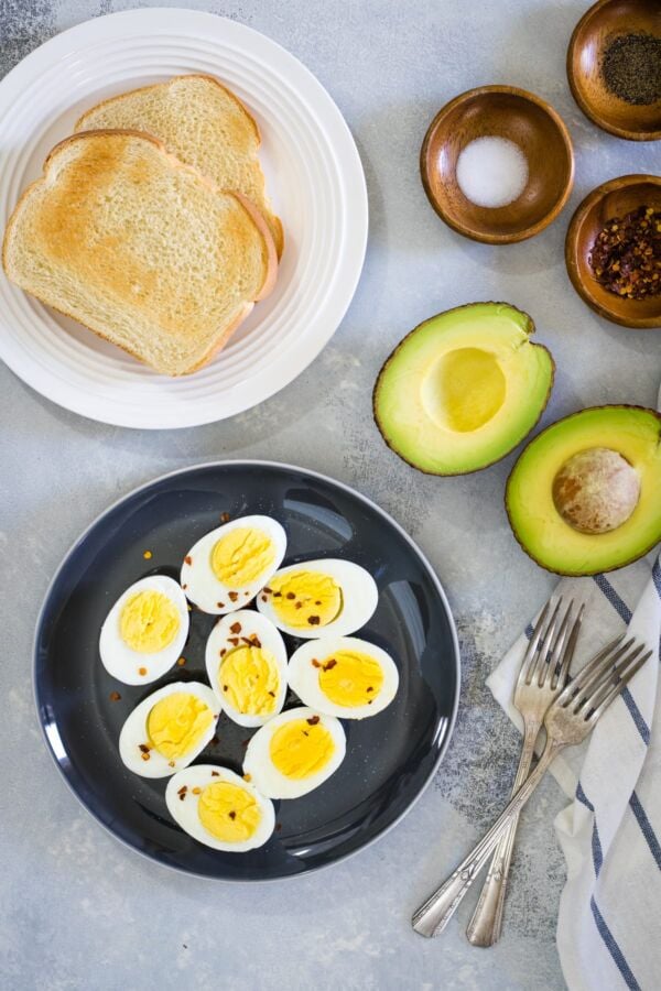 Hard boiled eggs on a black plate next to toast and avocado