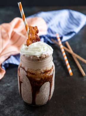 A bourbon milkshake in a mason jar topped with whipped cream and a piece of crispy bacon.