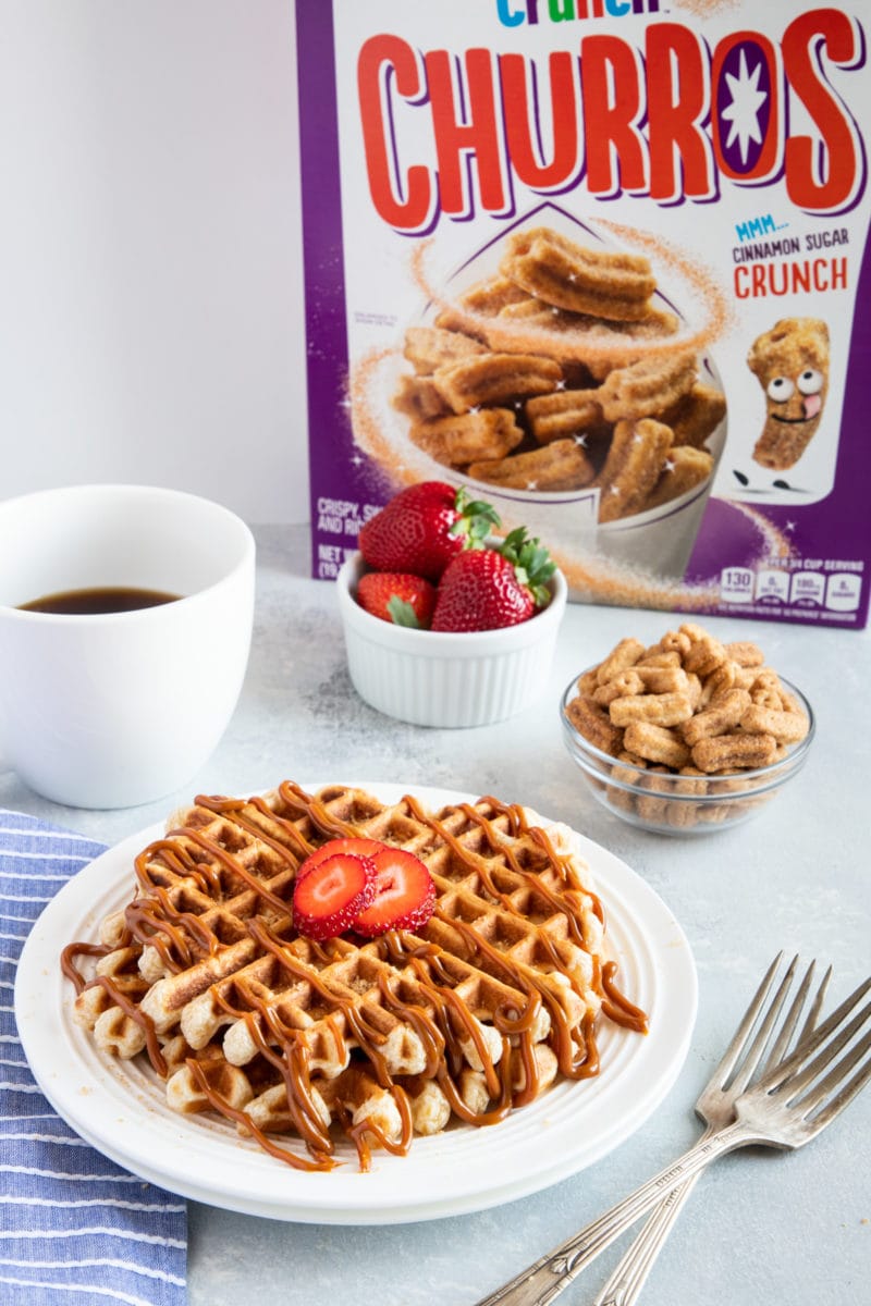 Waffles served on a white plate in front of fresh strawberries and a cereal box.