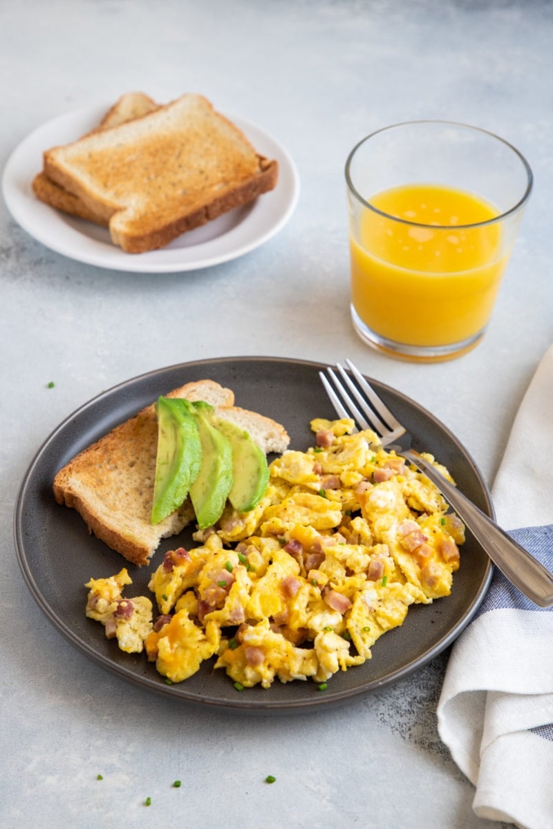 Ham and cheese scrambled eggs served on a plate with toast and avocado.