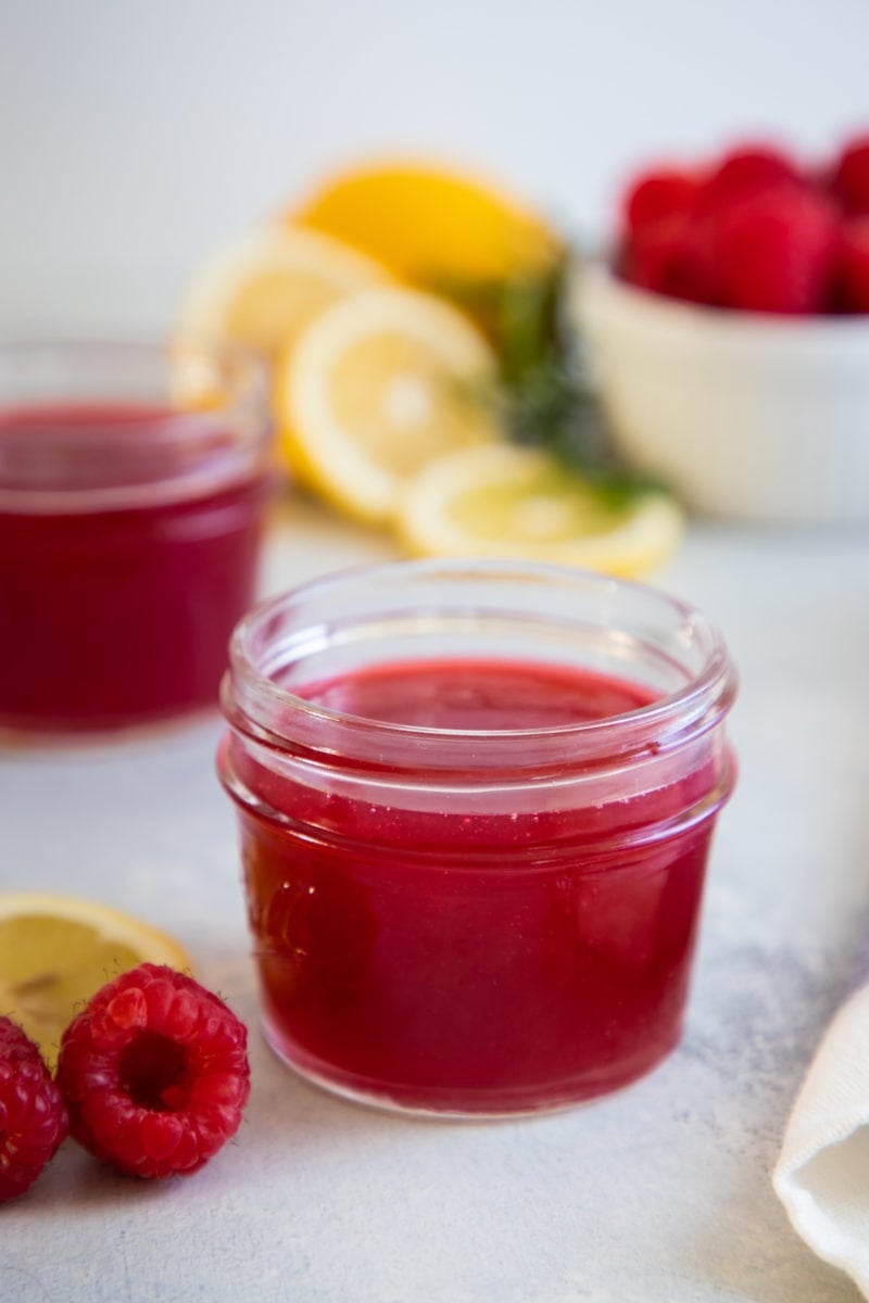 Close up of raspberry simple syrup in a small glass jar in front of lemon slices.