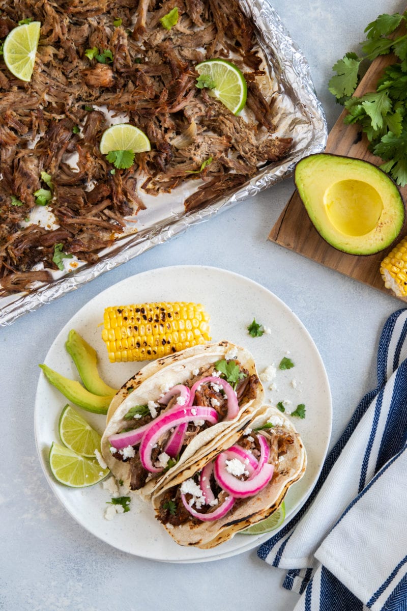 Pork Carnitas on a baking sheet and a plate with two served carnitas tacos  topped with pickled onions, avocado and corn. 