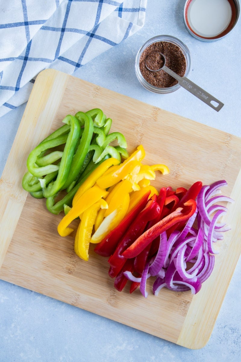 Red, green and yellow bell peppers cut into strips and sliced red onions on a wood board. 