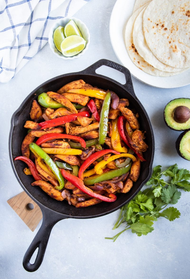 Chicken Fajitas in a skillet with flour tortillas, avocado, cilantro and lime wedges on the side. 