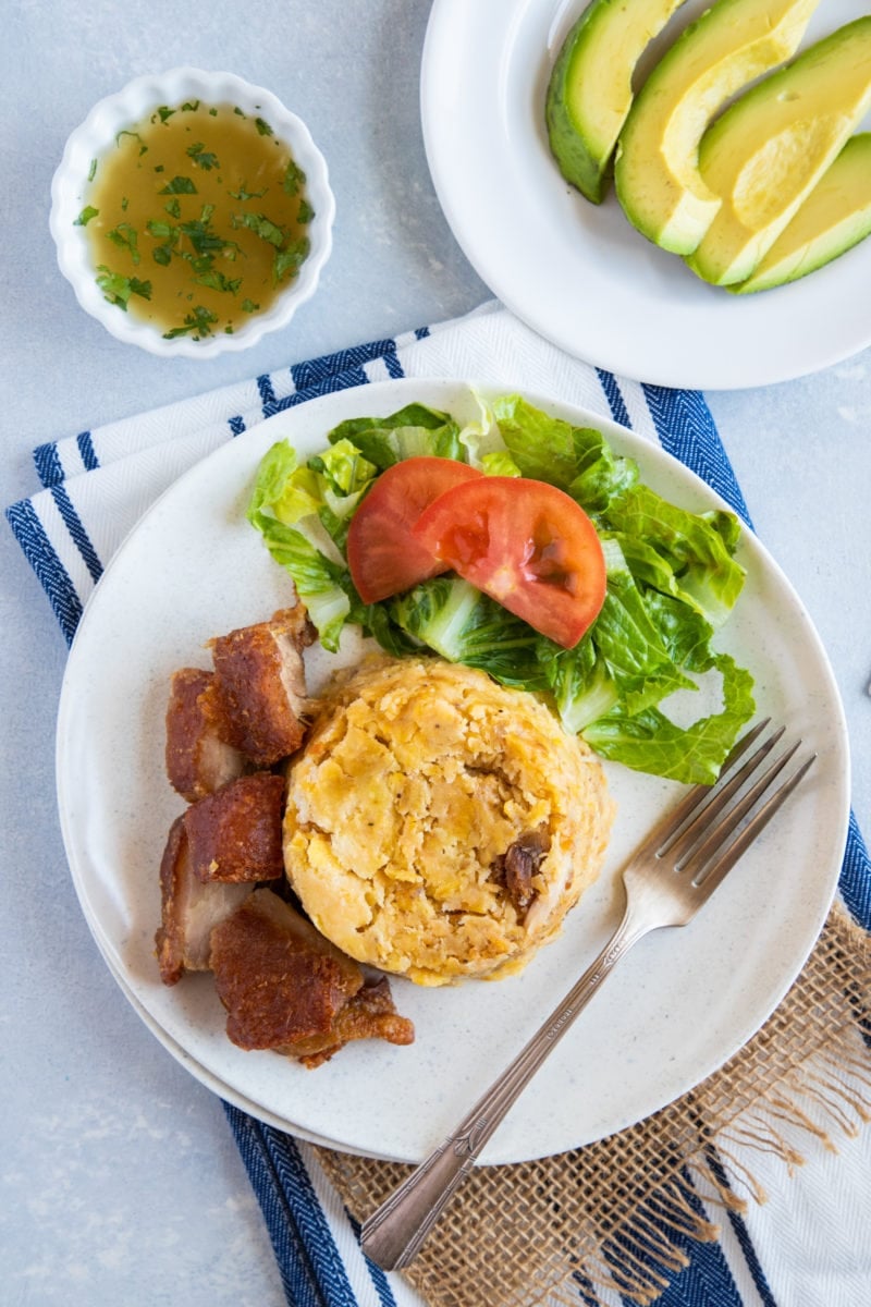 Traditional Mofongo served on a plate with extra chicharron and garlic mojo and avocado on the side. 