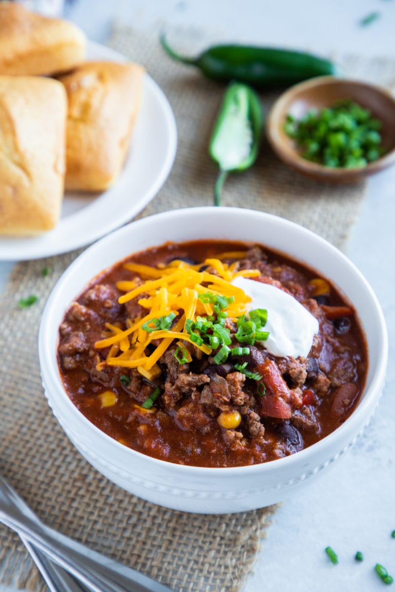 Slow cooker chili served in a white bowl topped with cheese, sour cream and scallions. Cornbread and jalapeño on the side. 