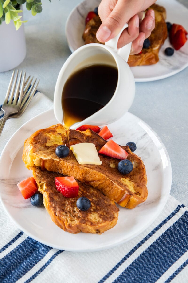Challah Bread French Toast on a plate topped with berries and hand pouring syrup