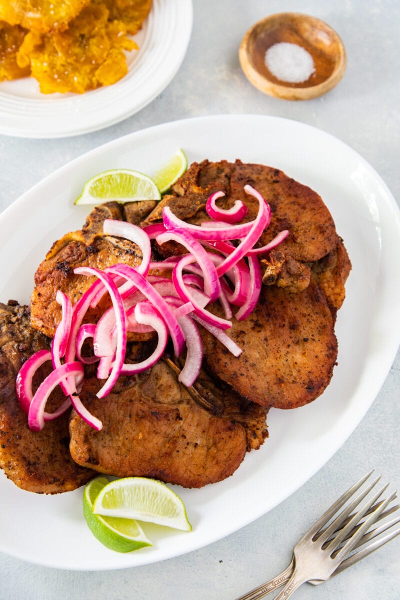 Dominican fried pork chops on a white plate served with lime wedges and red onion.