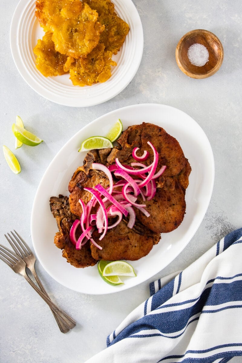 Overhead shot of Dominican fried pork chops on a plate next to a plate of platanos fritos.