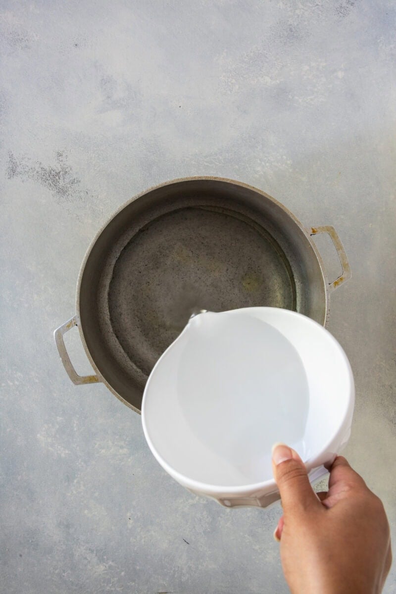 A hand pours water from a white measuring cup into a metal pot on a countertop, preparing for cooking.