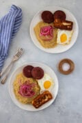 Two plates of mangĂș with fried eggs, salami, fried cheese, and pickled red onions.