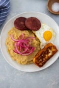 MangĂș with fried egg, salami, fried cheese, and pickled red onions on a white plate.