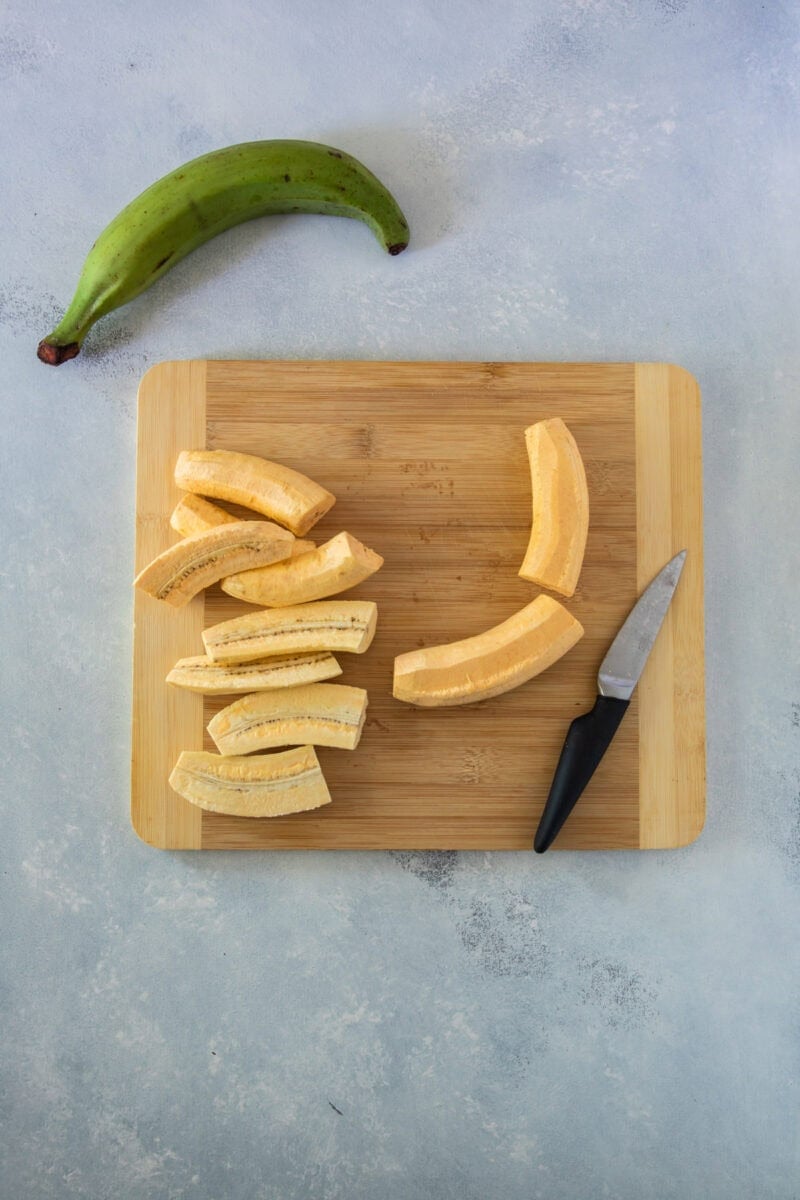 A peeled green plantain cut into chunks on a wooden cutting board with a knife, and an unpeeled plantain above it.