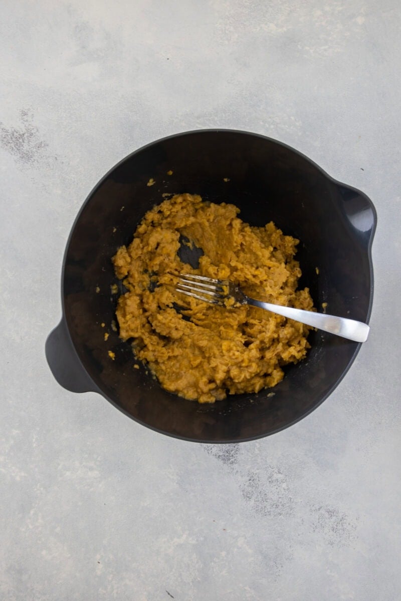 A black mixing bowl containing mashed plantains (mangĂș) with a fork inside, showing a soft, mashed texture.