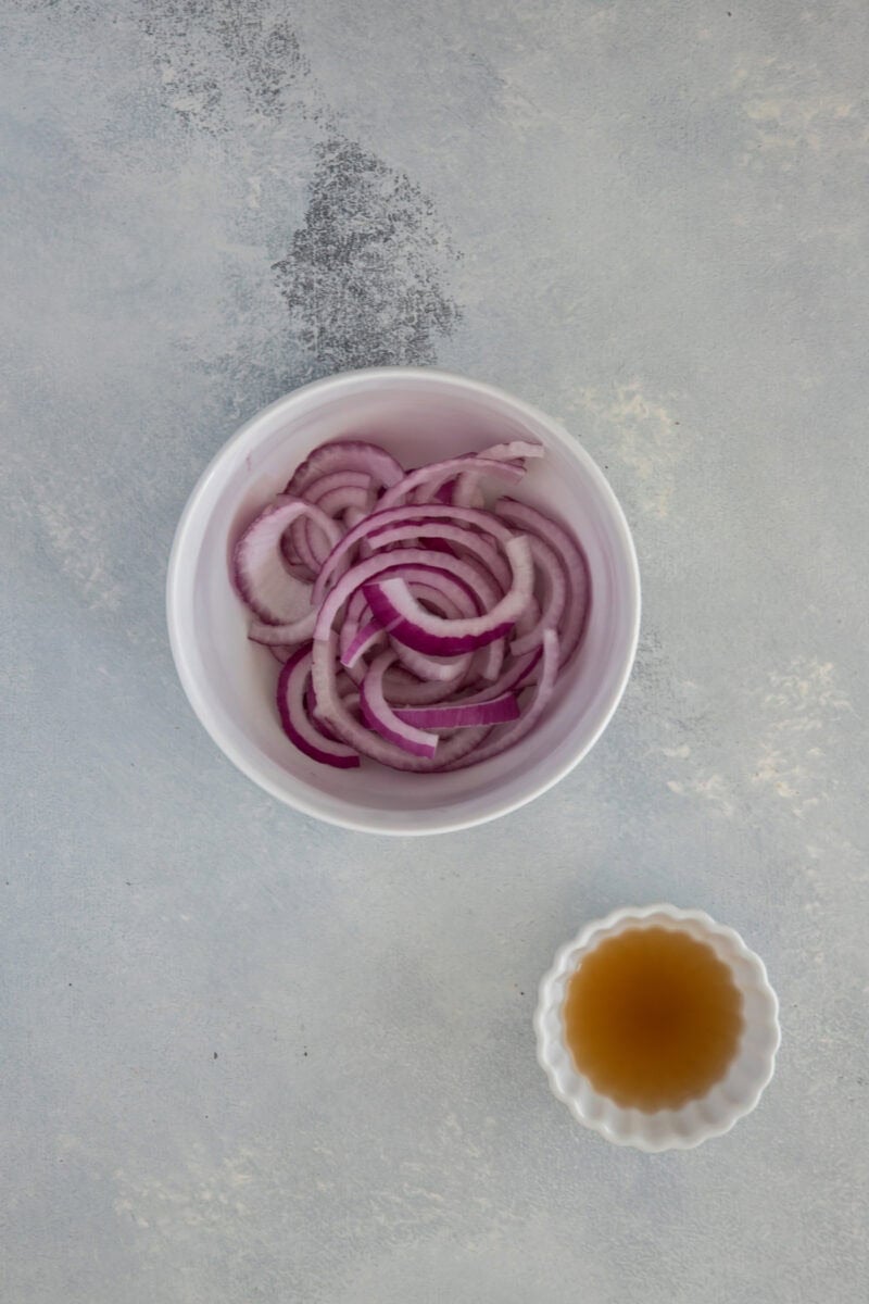 A white bowl filled with sliced red onions next to a small dish of liquid, likely vinegar or dressing.