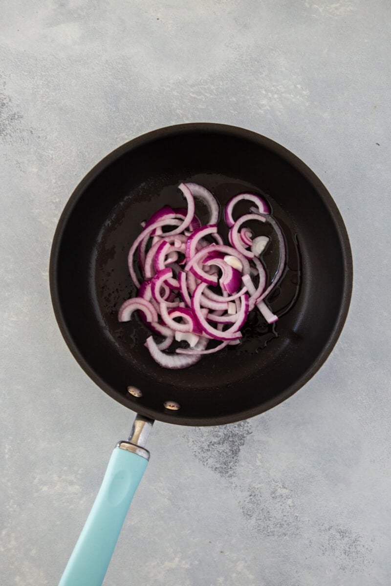 A black frying pan with sliced red onions cooking in oil on a light gray surface.