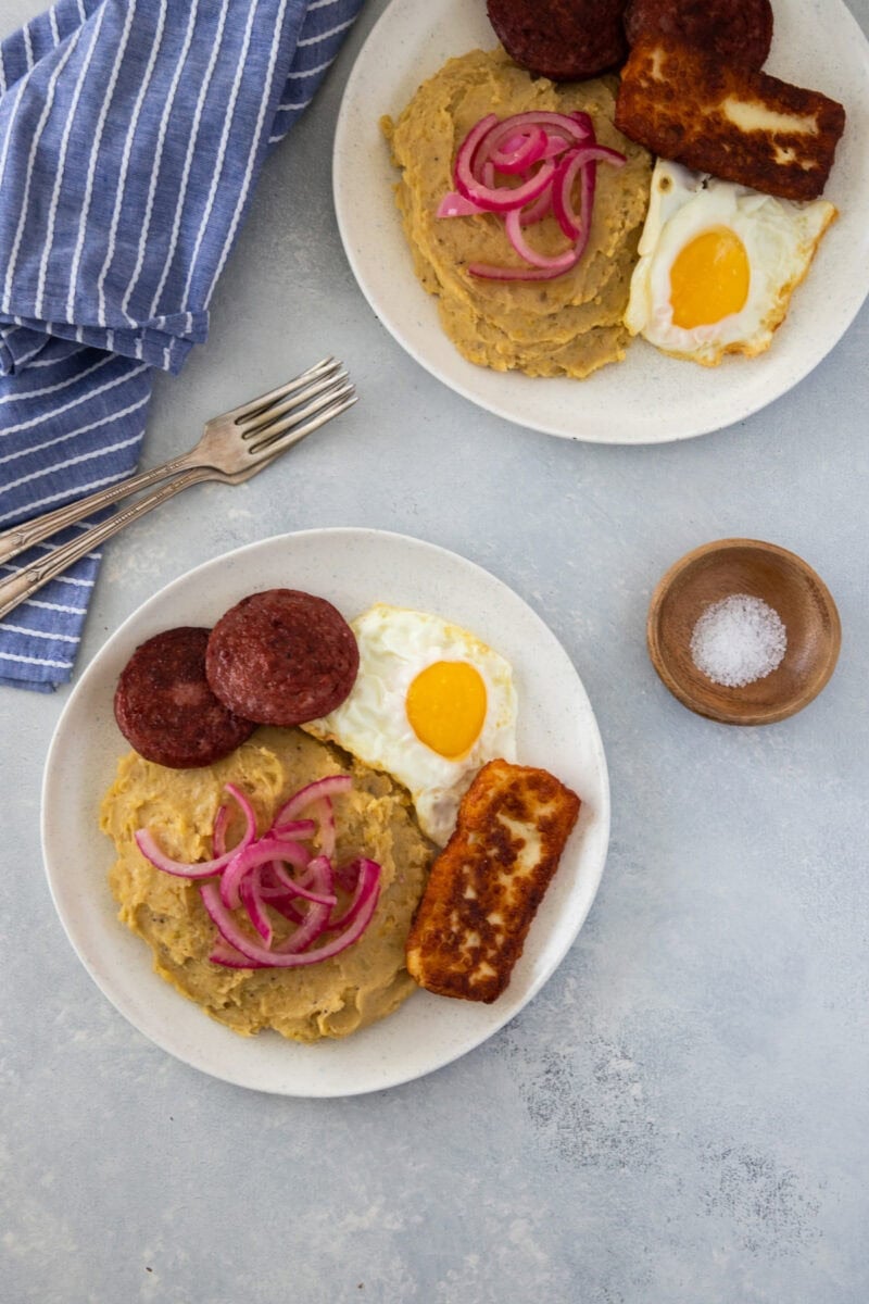Two plates of mangĂș with fried eggs, salami, fried cheese, and pickled red onions.