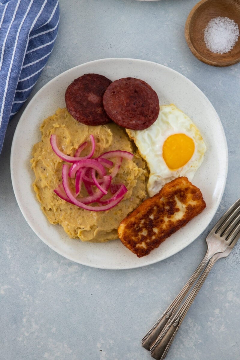 Mangú with fried egg, salami, fried cheese, and pickled red onions on a white plate.