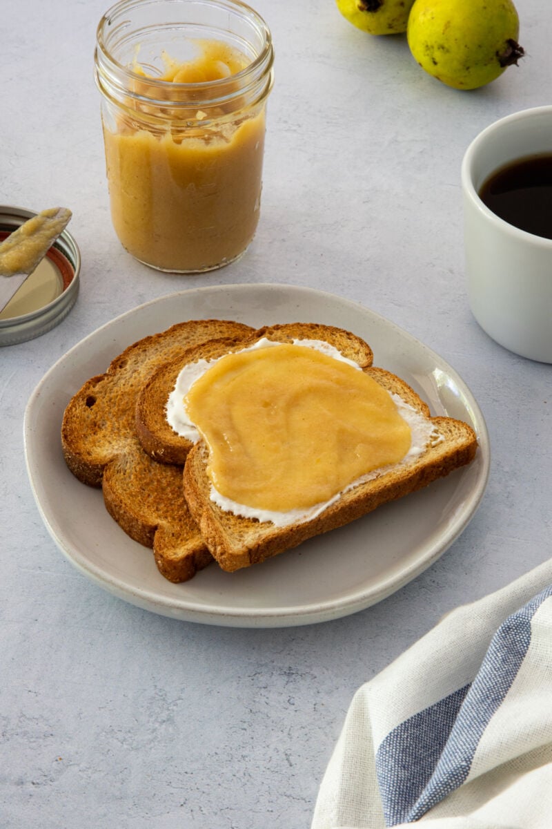 Tostadas de pan en un plato cubiertas con queso crema y mermelada de guayaba. Café al lado.