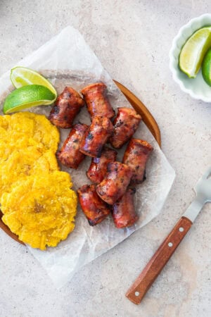 Longaniza Frita and fried plantains on parchment paper, served with lime wedges on a wooden board. Knife and bowl with lime slices on the side.