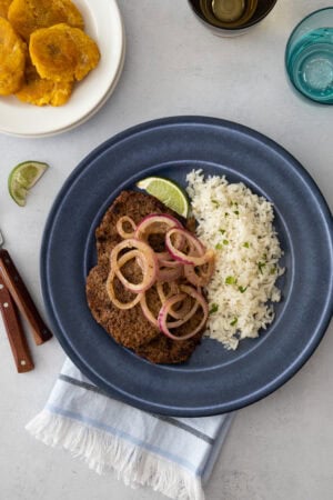A beef steak topped with onions, served with white rice and a lime wedge, ready to eat.