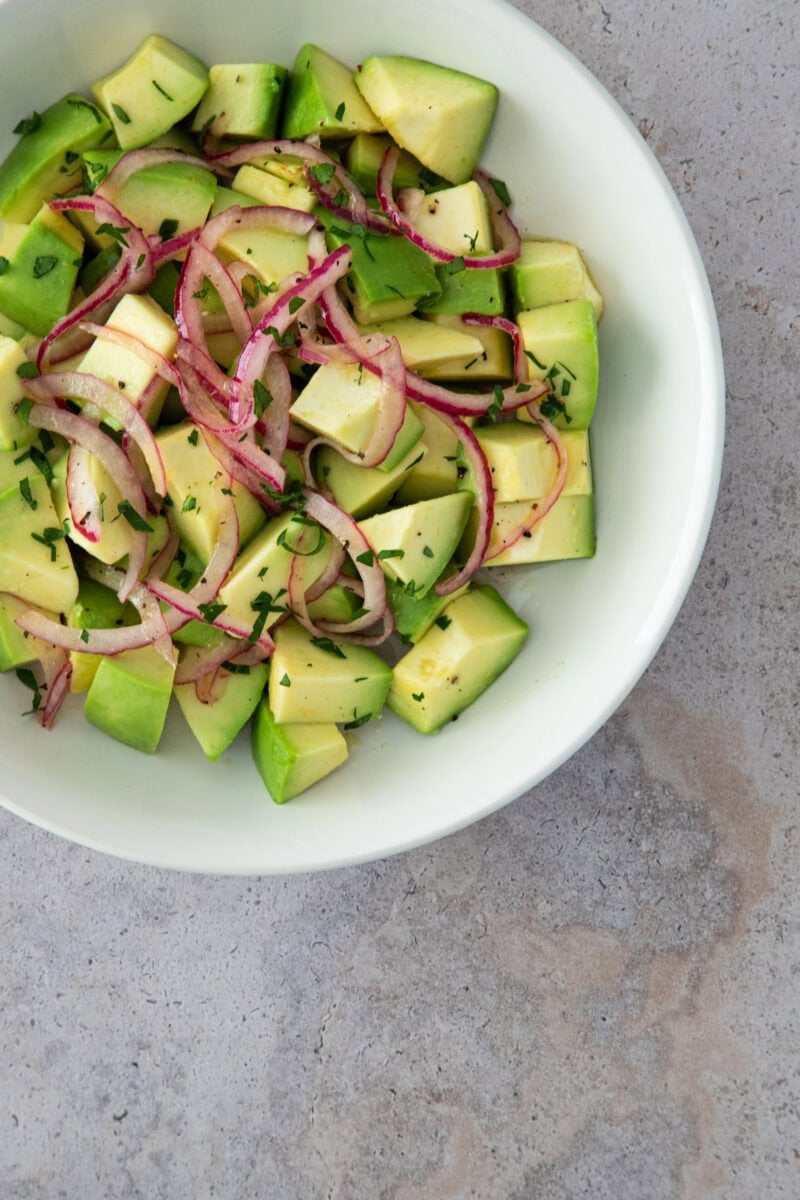 A fresh avocado salad with red onion slices and chopped herbs, served in a white bowl.