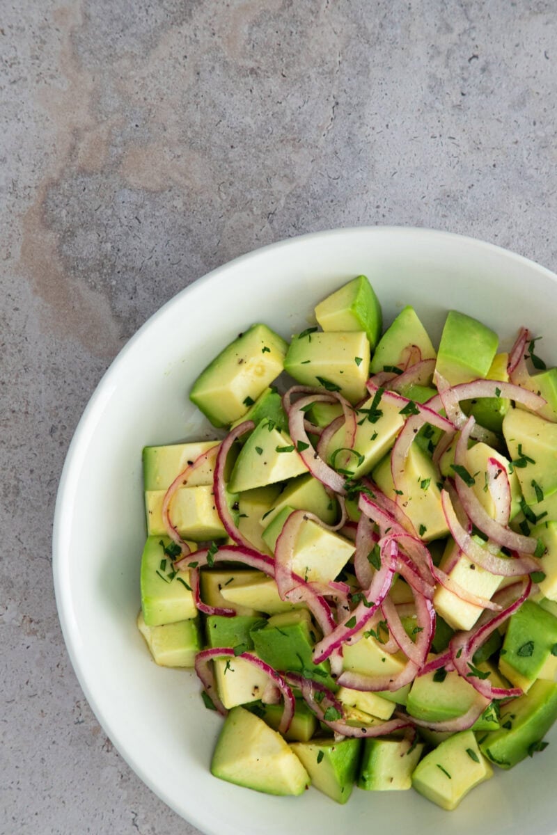 A fresh avocado salad with red onion slices and chopped herbs, served in a white bowl.
