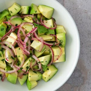 A fresh avocado salad with red onion slices and chopped herbs, served in a white bowl.