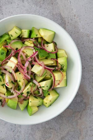 A fresh avocado salad with red onion slices and chopped herbs, served in a white bowl.