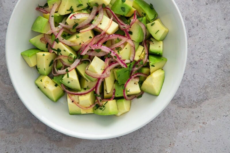 A fresh avocado salad with red onion slices and chopped herbs, served in a white bowl.