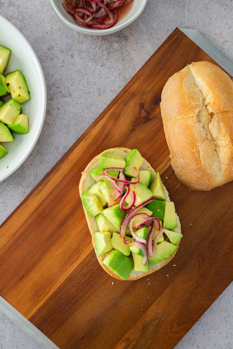 Tostada de aguacate con cebolla roja sobre pan rústico, servida en una tabla de madera.