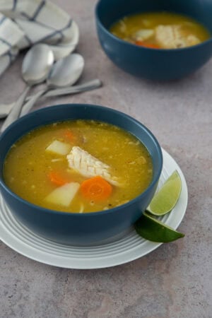 Dominican chicken soup with yuca, carrots, and tender chicken, served in a blue bowl with lime wedges.