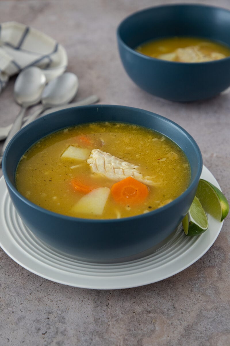 Dominican chicken soup with yuca, carrots, and tender chicken, served in a blue bowl with lime wedges.